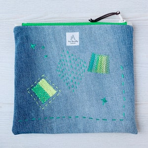 May include: A blue denim zippered pouch with green embroidery in geometric shapes. The pouch has a white label with the text "Anna Harwell Cadeaux".