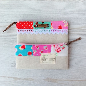 Può includere: Due pochette con cerniera in tessuto patchwork con un tiretto in pelle marrone. La pochette superiore ha un bordo in pizzo bianco e un'etichetta in legno con la scritta "Annie Thorntonley". La pochette inferiore ha una stampa floreale rosa, blu e verde e un'etichetta marrone con la scritta "travel is... This trip will stay with us and happy memory".