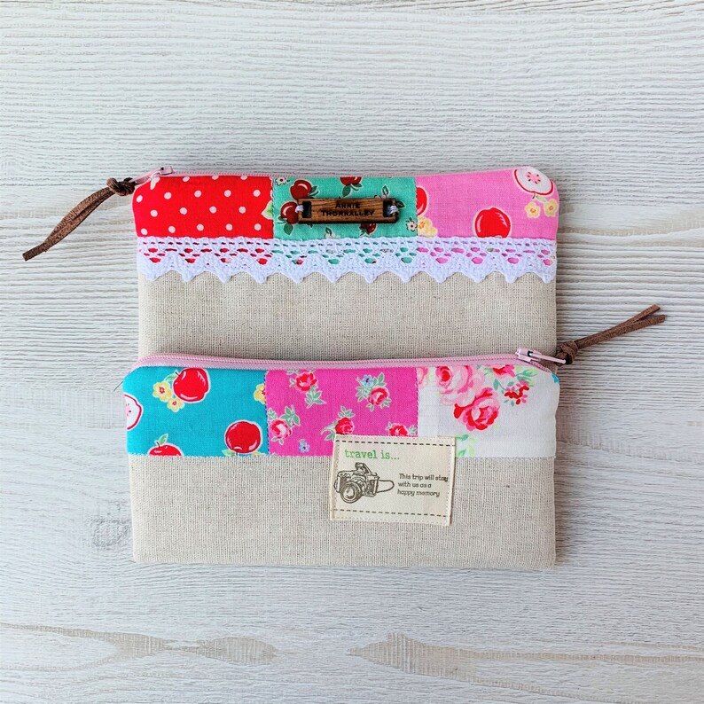 May include: Two patchwork fabric zippered pouches with a brown leather zipper pull. The top pouch has a white lace trim and a wooden tag that says "Annie Thorntonley". The bottom pouch has a pink, blue, and green floral print and a brown tag that says "travel is... This trip will stay with us and happy memory".
