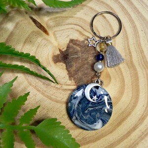 May include: A celestial-themed keychain featuring a round pendant with a blue and white marbled design, a silver crescent moon and star charm, and a silver tassel. The keychain includes a silver ring, beads, and a star charm.