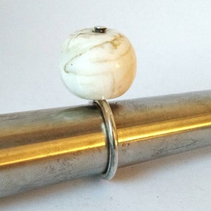 Könnte beinhalten: Ein weißer Ring mit einem runden, weißen, marmorierten Stein oben. Der Ring befindet sich auf einer silbernen Metallstange.