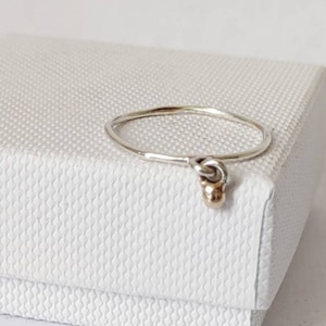 Op de afbeelding: Een zilveren ring met een kleine gouden kraal die eraan hangt. De ring ligt op een witte doos.