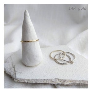 Peut inclure: Une collection de bagues délicates. Une fine bague en or martelé est présentée sur un support conique blanc. Trois bagues supplémentaires, dont une avec une texture perlée, sont disposées sur une surface blanche texturée. Le texte "14K gold" est visible.