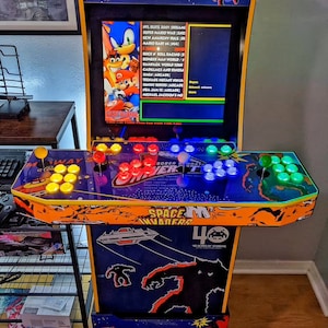 May include: A multi-colored arcade cabinet with a black screen displaying a list of games. The cabinet has a blue and orange design with the words "Space Invaders" and "40 Years of Space Invaders" printed on the front. The cabinet has a joystick and buttons with colorful lights.