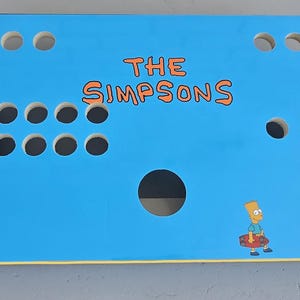 May include: A light blue arcade control panel with illustrations of The Simpsons characters. The panel has holes for buttons and joysticks, with Homer, Marge, Bart, and Lisa Simpson. The text "THE SIMPSONS" is in orange.