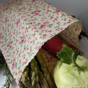 Könnte beinhalten: Wiederverwendbare Lebensmittelverpackung mit floralem Muster aus rosa Blüten und grünen Blättern auf cremefarbenem Hintergrund. Enthält frisches Gemüse wie Spargel, Radieschen und Kohlrabi.