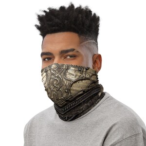 Puede incluir: Un hombre con una camisa gris y una m&aacute;scara facial con estampado negro y dorado. La m&aacute;scara tiene un dise&ntilde;o que se asemeja a una armadura.