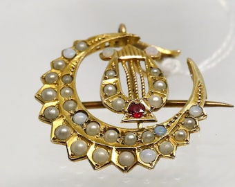 May include: A gold crescent moon brooch featuring a central lyre motif. The crescent is decorated with a series of small white pearls along its outer edge. A single deep red gemstone is set at the base of the lyre, which is also accented with pearls.