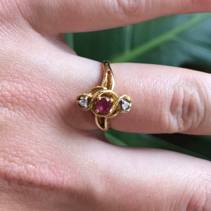 May include: A gold ring with a red gemstone and two smaller white gemstones. The ring is set in a twisted band.
