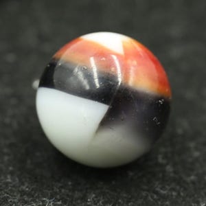 May include: A colorful glass marble with a mix of white, black, orange, and red sections. The marble has a glossy finish and is set against a dark background. This collectible toy is a classic.