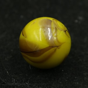 May include: A yellow glass marble with brown marbling. The spherical marble has a glossy finish and is set against a dark background. The classic design features swirling patterns.