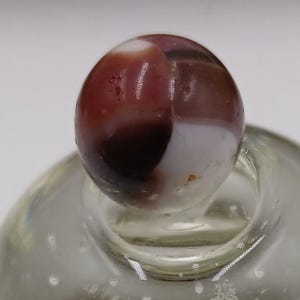 May include: A clear glass bottle with a round body and a decorative stopper. The stopper is a marble with a mix of brown, white, and red colours. The bottle is filled with a clear liquid.