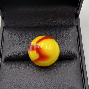 May include: A yellow glass marble with red and orange swirls, presented in a black jewellery box. The marble is spherical and has a glossy finish, with the red and orange patterns creating a striking visual contrast against the yellow base.