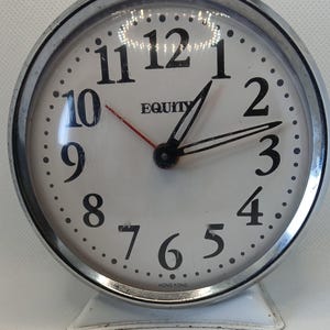May include: A silver alarm clock with a white face and black numbers. The clock has a red second hand and black hour and minute hands. The clock face says "EQUITY" and "HONG KONG".