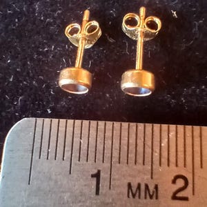 May include: A pair of gold stud earrings with small, round, blue gemstones. The earrings are measured against a ruler, showing the gemstones are approximately 2 millimeters in diameter.