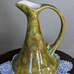 May include: A green ceramic jug with a white interior and a distinctive, curved handle. The jug has a mottled glaze with brown and green accents.