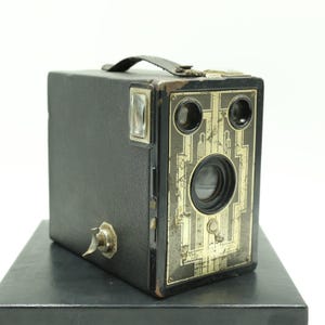 以下が含まれることがあります： 装飾的な金色の前面プレートが付いた、ヴィンテージの黒いボックスカメラ。 カメラには革製のハンドルとフラッシュアタッチメントが付いています。 レンズは前面の中央に配置され、その上に2つの小さなレンズがあります。 カメラは黒い表面に置かれています。