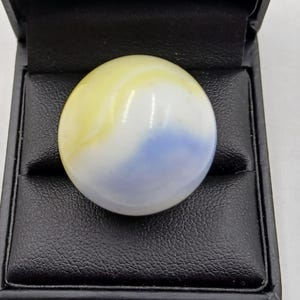 May include: A large, round marble with a swirl of yellow, white, and blue colours. The marble is set in a black, leather-like box, creating a striking contrast. The marble's polished surface reflects light, highlighting its unique colour pattern.