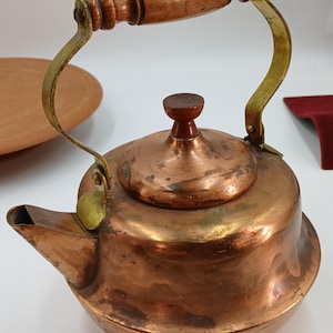 May include: A copper kettle with a wooden handle and lid knob. The handle is attached with brass brackets. The spout is on the left side of the kettle. The kettle has a round body and a slightly tarnished finish.