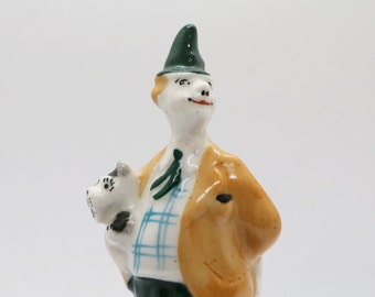 May include: A ceramic figurine of a man in a green hat and tan jacket, holding a small dog. The man has a white shirt with a blue plaid vest and a green tie. The dog is white with black spots.