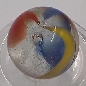 May include: A clear glass marble with red, blue, and yellow swirls. The marble has a few small imperfections and is set in a clear glass holder. The colors are vibrant and the design is eye-catching.