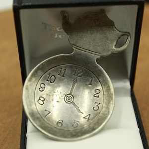 Peut inclure: Un montre de poche argentée avec un design de théière sur le dessus. Le cadran de la montre affiche les chiffres de 1 à 12, avec les aiguilles pointant vers environ 4 heures. La montre est présentée dans un coffret noir avec un intérieur blanc.