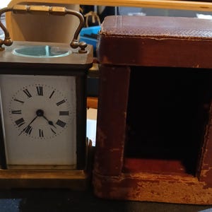 May include: A rectangular brass carriage clock with a white face, black Roman numerals, and a top handle, positioned next to its matching brown leather-covered protective carrying case with a hinged lid and visible wear on the edges.