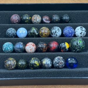 Peut inclure: Une collection de vingt-et-un billes de verre colorées, présentées dans un plateau noir. Les billes présentent divers motifs et couleurs, notamment des motifs mouchetés, tourbillonnés et unis. Les couleurs incluent le rouge, le bleu, le vert et le jaune.