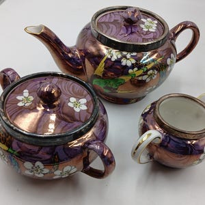 May include: A three-piece tea set with a purple and bronze metallic finish. The set includes a teapot, a sugar bowl with a lid, and a creamer. Each piece is decorated with white floral accents and a bird design.