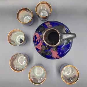 May include: A ceramic sake set. The carafe is round, blue and black with a handle. Six matching cups surround the carafe. The carafe and cups have gold rims and a red and pink floral design.