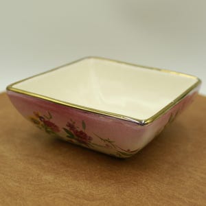 May include: A square ceramic bowl with a cream interior and a pink exterior decorated with floral patterns. The rim is trimmed with gold. The bowl is on a wooden surface.