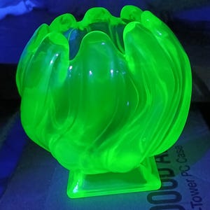 May include: A bright, neon green glass bowl with an unusual, flowing form. The bowl features a flared, petal-like edge and rests on a square base. The glass has a swirling, textured design throughout, giving it a unique appearance.