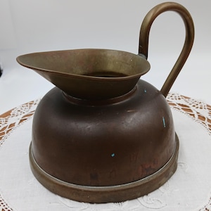 May include: A vintage copper pitcher with a wide, rounded base and a prominent, curved handle. The metal surface displays a weathered patina with subtle oxidation marks, featuring a wide, open spout and a smooth, circular rim at the top.
