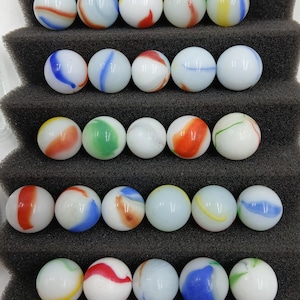 May include: A collection of twenty-five spherical glass marbles arranged in five rows on a dark gray foam display stand. Each marble features a white opaque base with various colorful swirls in shades of red, blue, green, and yellow.