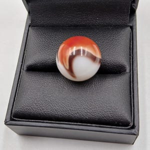 May include: A vibrant marble with a red, brown, and white swirl design, presented in a black display box. The marble is spherical and has a polished surface, highlighting the detailed pattern. The box is open, displaying the marble.