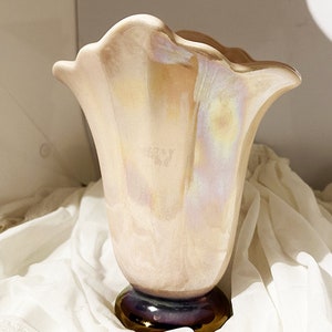 May include: A pale pink ceramic vase with an iridescent glaze. The vase has a wide, flared opening and a narrow base. The base is painted with a dark purple and gold design.