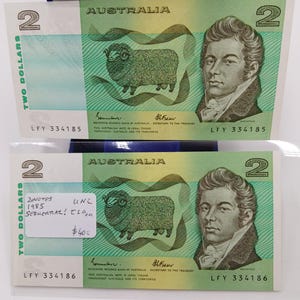 May include: Two Australian two-dollar banknotes. The notes are green with a portrait of a man on the right and a sheep on the left. The text "AUSTRALIA" and "TWO DOLLARS" are visible. One note has a sticker with text.