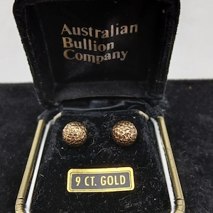 May include: A pair of round, textured gold stud earrings presented in a black velvet jewellery box. The interior lid features the text Australian Bullion Company, and a small rectangular label below the earrings reads 9 CT. GOLD.