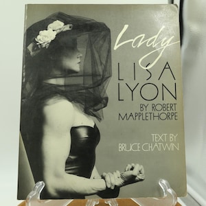 May include: A vintage book cover featuring a black and white photograph of a person wearing a black veil and a strapless top. The title "Lady Lisa Lyon" is in white script, with author and text credits.