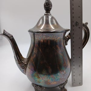 May include: A silver teapot with an ornate design and three feet. The teapot has a lid with a finial and a handle with a decorative design.