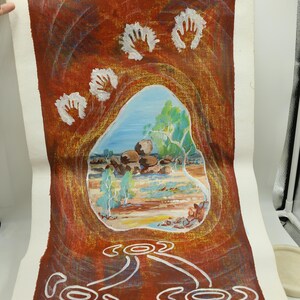 May include: An abstract painting on canvas featuring a landscape scene within a cave-like structure. The artwork uses warm tones of red and brown, with white handprints and stylized symbols. The landscape depicts rocks, trees, and a blue sky.