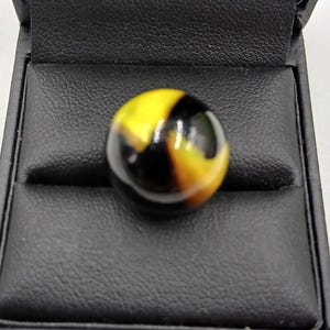 May include: A yellow, black, and amber marble is displayed in a black, leather-effect box. The marble is spherical and has a glossy finish. The box is open, showcasing the marble.