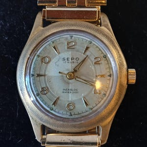 May include: A gold-toned SEPO wristwatch with a rectangular link band. The watch face is light-colored with black numerals and hands. The watch face reads "SEPO 17 RUBIS INCABLOC SWISS MOV".