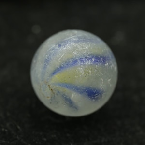 May include: A close-up of a clear glass marble with blue and yellow swirls. The marble is round and has a slightly frosted appearance. The background is a dark, solid color.