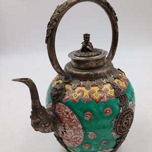 May include: A round ceramic teapot featuring a vibrant green base with red and yellow decorative patterns. The teapot is encased in ornate silver-toned metalwork, including a curved handle with a red bead, a spout, and a lid topped with a small figure.