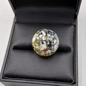 May include: A speckled, spherical stone, mainly black, white, and yellow, sits in a black jewellery box. The stone's surface has a mottled appearance, with various colours and textures. The box is open, revealing a soft, black interior.