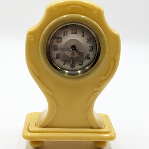 以下が含まれることがあります： 丸い文字盤とローマ数字の小さなクリーム色の装飾時計。時計は長方形のベースを持ち、角が丸くなっています。