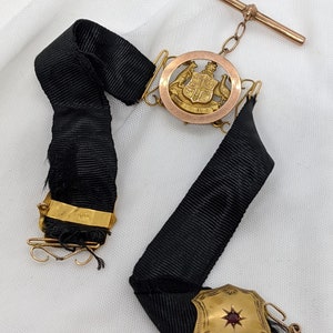 May include: A gold and black ribbon with a gold pendant featuring the Australian coat of arms. The pendant is attached to a gold chain and a gold bar. The ribbon has a gold clasp with a red stone.