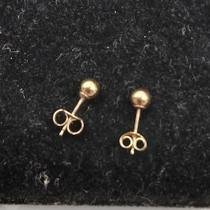 May include: A pair of gold-toned ball stud earrings featuring a smooth, spherical design at the front and a secure butterfly-style backing post. These earrings have a polished metallic finish and are presented as a matching set of two individual pieces.