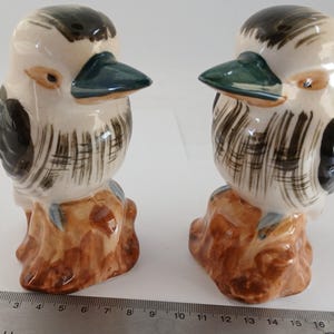May include: Two ceramic kookaburra bird figurines, each perched on a brown tree stump. The birds are painted with brown, black, and green details.
